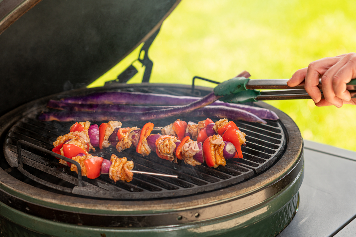 Kamado Griller mit Gemuesespieß und Melanzani auf dem Rost des gruenen kamadogrillers und eine Zange die mit einer Hand bedient wird